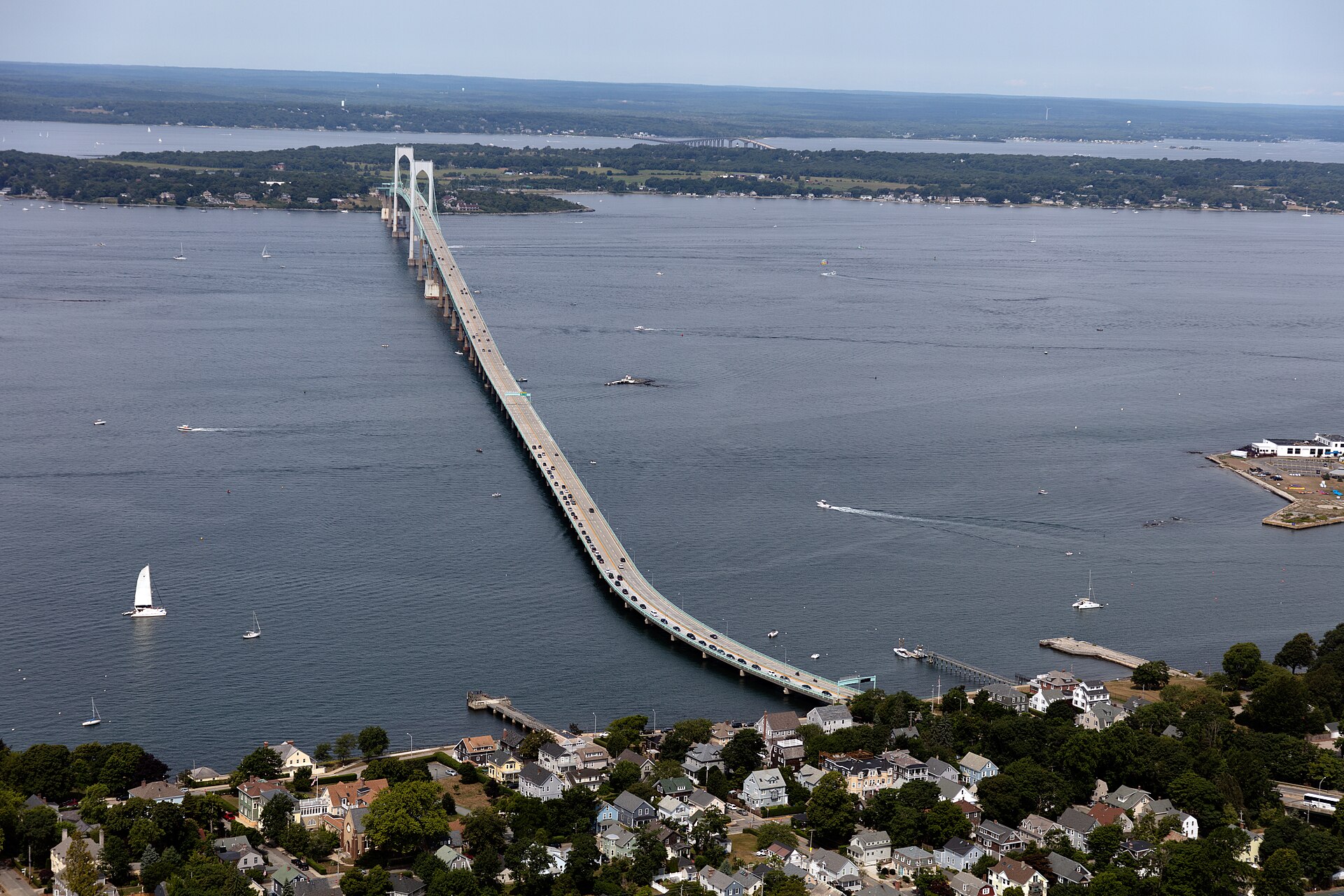 Aerial photograph of the Claiborne Pell Bridge, also called the Newport Bridge, crossing Narragansett Bay toward Newport, with sailboats on the water and houses along the near shore.